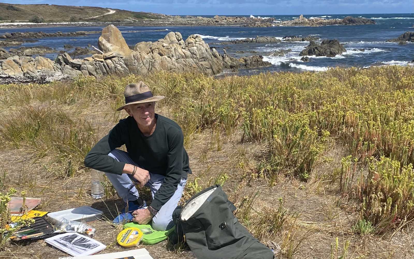 mike-staniford-artist-australia2 A person wearing a hat and dark long-sleeved shirt sits on grass near the rocky coastline, surrounded by art materials, paper, and a rucksack—capturing their inspiration much like the creative process described in About Mike Staniford. The sea and rocks are visible in the background under a blue sky.