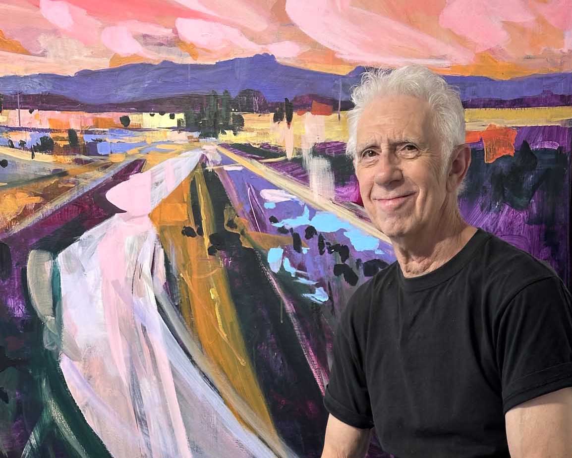 An older man with white hair, wearing a black T-shirt, sits in front of a colourful abstract painting featuring a landscape with a road, fields, mountains, and a vibrant pink and orange sky—an inspiring backdrop that reflects the creative spirit of Mike Staniford. Join our mailing list to stay updated on his latest works and exhibitions.